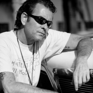 Photographer Neil Zlozower in sunglasses and white tee, with arm rested on hood of white car.