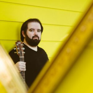 Brian Wolff with acoustic guitar on yellow backdrop