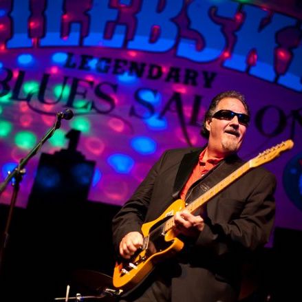 Reverend Raven playing electric guitar on colorfully lit stage.