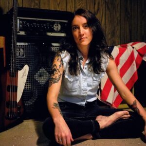 Sylvia Rose Novak sitting in front of her amp, a bass guitar and American flag