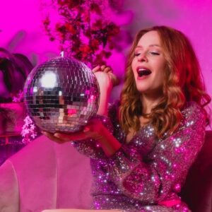 Hiedi Webster looking at a mirror ball, in glamorous attire on pink background