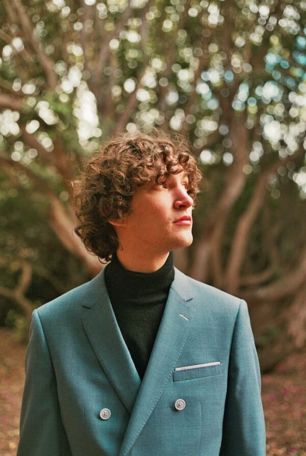 Otto standing outdoors in wooded backdrop, wearing a teal sports coat and black turtleneck sweater, looking updward