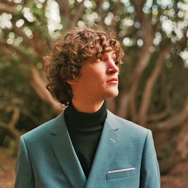Otto standing outdoors in wooded backdrop, wearing a teal sports coat and black turtleneck sweater, looking updward