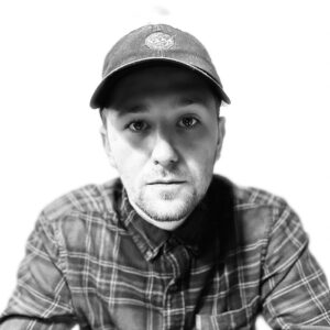 Andrew Chris, black & white photo, wearing ball cap and a flannel shirt on a white background