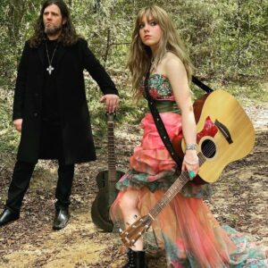 Cozy Anda Flounder, Cozy and Ezra Vancil with guitars standing in wooded area. Ezra in black, Cozy in colorful dress.