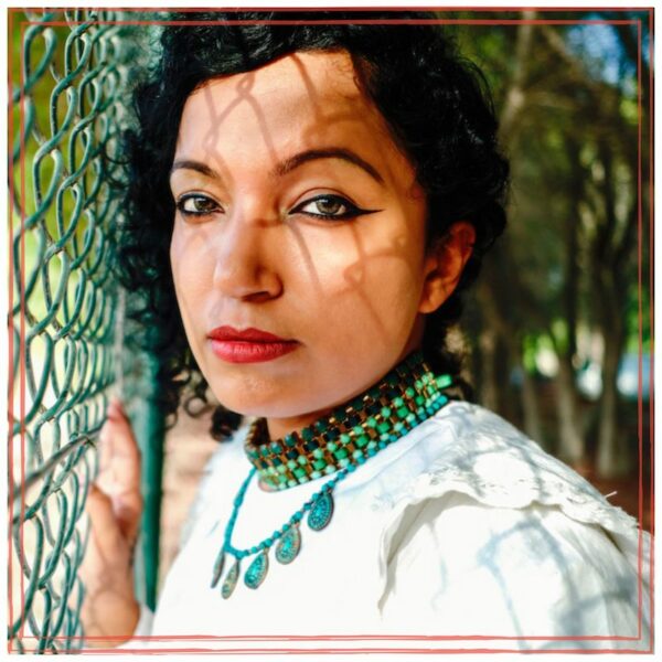 Shilpa Ananth with hand on green cyclone fence, casting shadows on her face. Wearing white top.