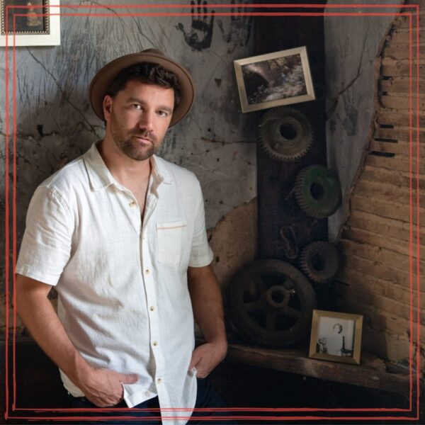 Eli Lev standing in room of an old building, wearing white shirt wearing a hat with hands in pocket. | Artwork for Unstarving Musician episode 332 Eli Lev – Spiritual Growth: From 250 Shows to Finding Sacred Space in Music