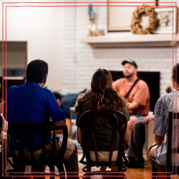 Acoustic musician performing in a living room for a small seated audience, with a fireplace and home decor visible in the background — a typical house concert setting.