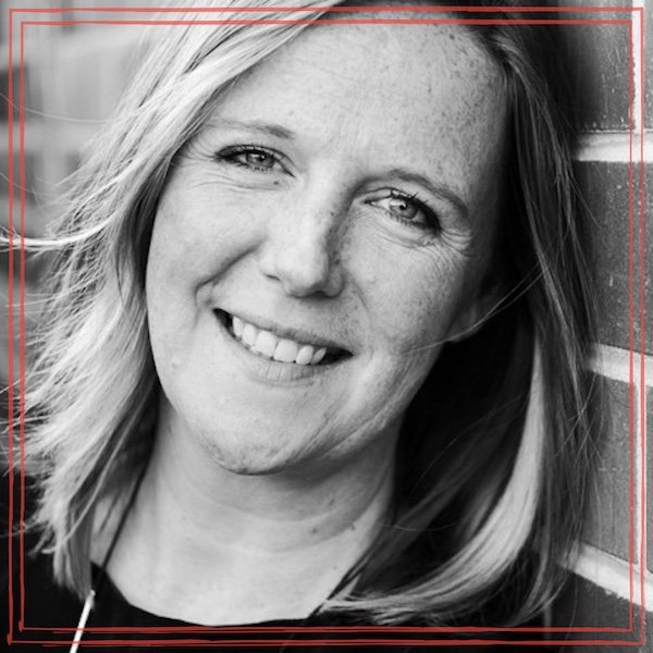 Black and white headshot of Lynz Crichton, smiling with head tilted next to a brick wall