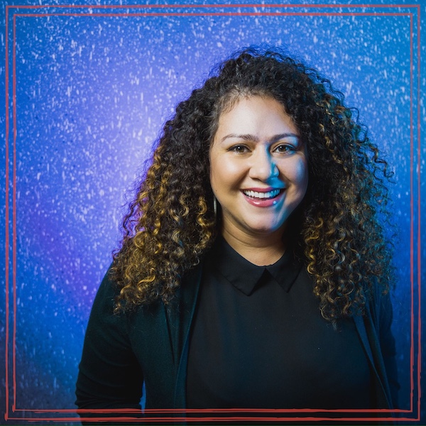 Janette Berrios smiling, with long curly hair, wearing a black collared top against a blue and purple backdrop.