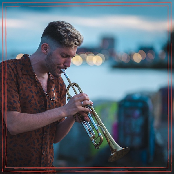 Noam Bargil playing trumpet outdoors at dusk in a brown patterned shirt, with blurred city lights across the water behind him and a thin red border around the photo.